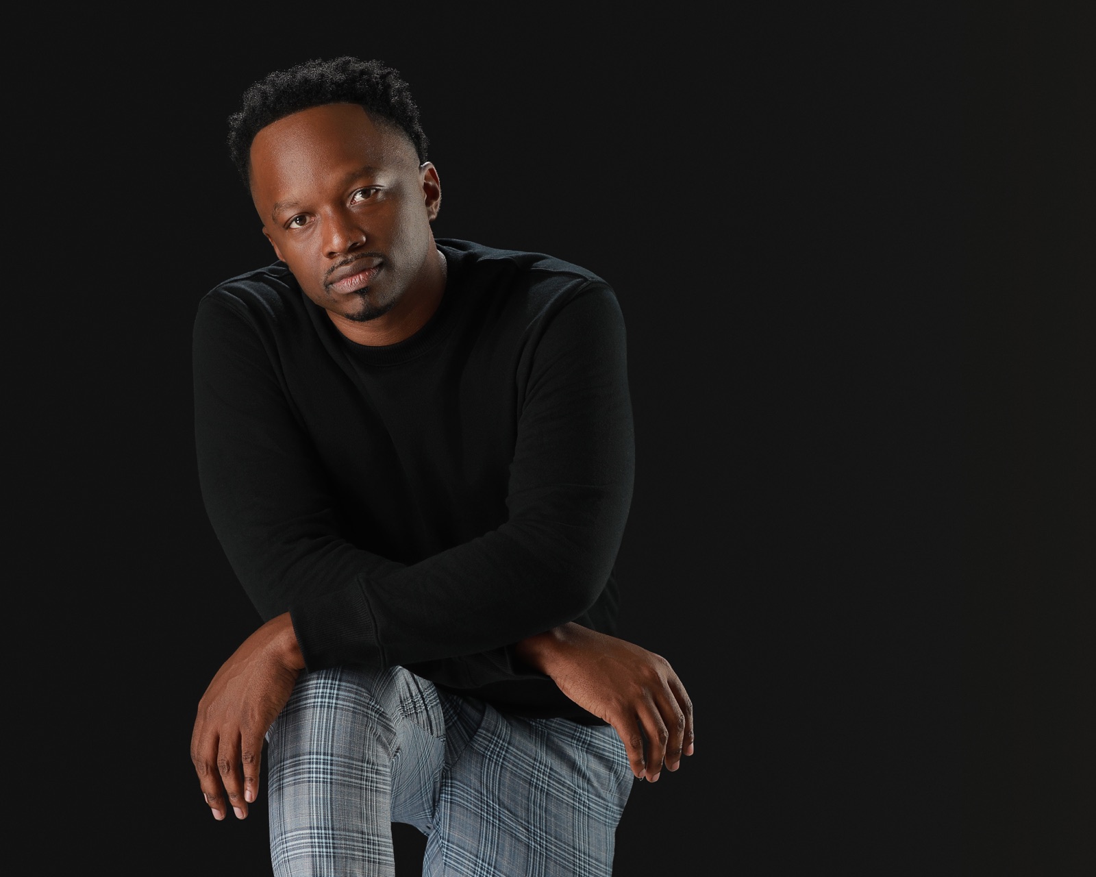Brian D. Washington in a black sweatshirt with crossed arms and plaid trousers, photographed against black